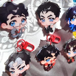 Puede incluir: Una colección de seis llaveros de acrílico con personajes estilo chibi de cabello oscuro y ojos azules. Cada dije incluye un broche de langosta de plástico de colores. Los personajes lucen diversos atuendos, incluyendo trajes y ropa casual, con rasgos faciales expresivos.