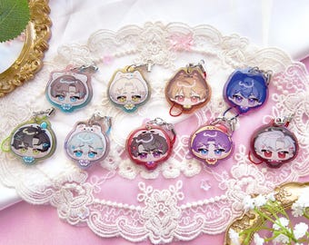 May include: A collection of anime-style character keychains. Each keychain features a unique character design with different hair colors and outfits. The keychains are attached to silver-colored metal clasps and are displayed on a pink lace surface.