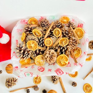 Puede incluir: Un tazón blanco lleno de rodajas de naranja secas, palitos de canela y piñas. El tazón está sobre una superficie blanca con las palabras "Merry Christmas" en letras rojas arriba.