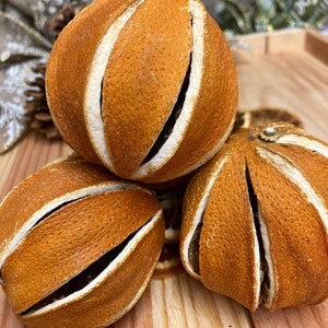 May include: Three dried orange slices, each with a unique pattern of cuts, are arranged on a wooden surface. The orange slices are a deep orange color and have a slightly textured surface.