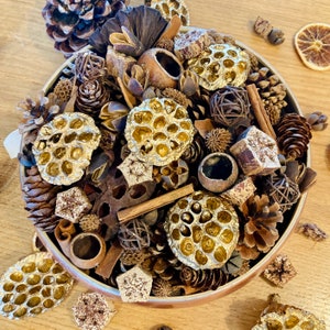 May include: A decorative bowl filled with a variety of natural potpourri elements. The arrangement includes pine cones, dried seed pods, cinnamon sticks, and other dried botanicals in shades of brown, gold, and beige. The bowl is a light brown colour.