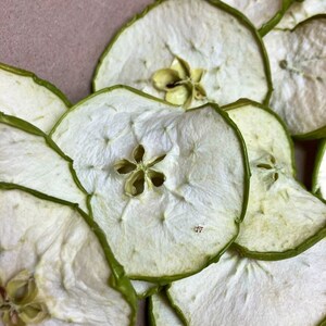 May include: A close-up of a pile of dried apple slices. The apple slices are light brown in color and have a green rim. The slices are arranged in a circular pattern.