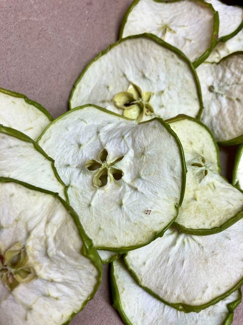 May include: A close-up of a pile of dried apple slices. The apple slices are light brown in color and have a green rim. The slices are arranged in a circular pattern.