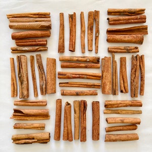 May include: A collection of cinnamon sticks arranged in a grid pattern on a white surface. The sticks are various lengths and thicknesses, with a natural brown color.