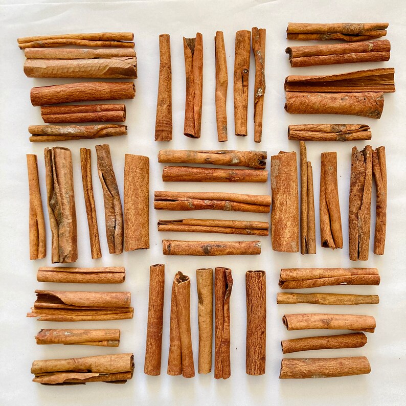 May include: A collection of cinnamon sticks arranged in a grid pattern on a white surface. The sticks are various lengths and thicknesses, with a natural brown color.