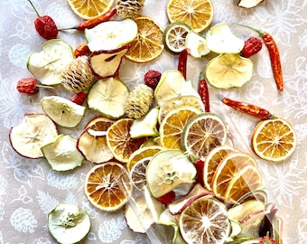Könnte beinhalten: Ein klarer Plastikbeutel, der mit getrockneten Fruchtscheiben überquillt, darunter Orangen, Äpfel und Limetten. Rote Chilischoten und kleine Tannenzapfen sind auf einer gemusterten Oberfläche um den Beutel herum verstreut. Die Fruchtscheiben sind in verschiedenen Braun- und Gelbtönen gehalten.