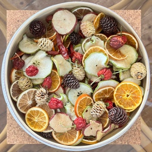 Op de afbeelding: Een kom gevuld met gedroogde fruitschijfjes, dennenappels en rode pepers. De fruitschijfjes zijn een mix van appels, sinaasappels en peren. De dennenappels zijn bruin en hebben een natuurlijke, rustieke uitstraling. De pepers zijn rood en voegen een vleugje kleur toe aan de kom.
