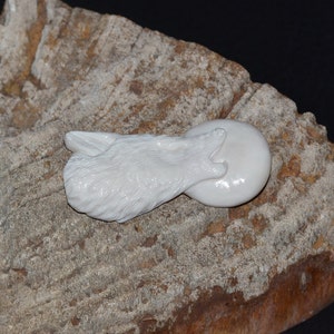 May include: A white carved wolf head pendant with a smooth, round, white stone. The pendant is resting on a rough, brown, textured surface.