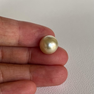 Könnte beinhalten: Eine einzelne, goldfarbene Perle. Die runde Perle hat eine glatte, reflektierende Oberfläche. Sie wird zwischen zwei Fingern gehalten, was ihre Größe und Farbe hervorhebt. Das Bild zeigt die natürliche Schönheit der Perle, ein klassisches Schmuckelement.