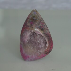 May include: A pink and gray agate cabochon with a crystal-lined cavity. The agate is polished and has a smooth, rounded shape.