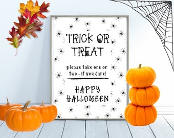 May include: A white sign with black spider illustrations and the text "Trick or Treat, please take one or two - if you dare! Happy Halloween".  There are three orange pumpkins stacked on a white surface.