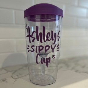 Puede incluir: Un vaso de plástico transparente con una tapa morada y un diseño de vinilo morado que dice "Ashley's Sippy Cup" con un corazón.