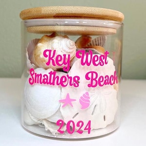 May include: A clear glass jar filled with seashells, topped with a bamboo lid. The jar has pink text that reads "Key West Smathers Beach 2024" with star and seashell graphics. The jar is filled with various white and tan seashells.