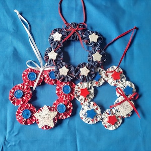 May include: Three handmade fabric wreaths in red, white, and blue with star and button embellishments. The wreaths are decorated with the word "USA" on two of them.