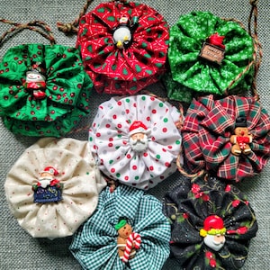 May include: Nine handmade Christmas ornaments in various colors and patterns. Each ornament features a different festive character, including Santa Claus, a penguin, a snowman, a gingerbread man, and a reindeer. The ornaments are made of fabric and have a ruffled appearance.