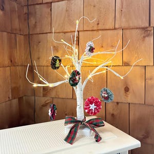 May include: A white birch tree with lights and fabric ornaments in red, green, and white. The tree is on a white table with a red and green plaid bow.