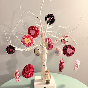 May include: A decorative white birch tree with hanging fabric ornaments in various colors and patterns. Some ornaments have text that reads "HUG ME", "SAY YES", and "BE KIND". A small owl figurine sits at the base.