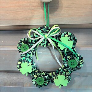 May include: A St. Patrick's Day wreath with a black fabric base covered in green shamrocks. The wreath is adorned with green shamrock cutouts, green buttons, and a green and white ribbon bow. A green ribbon is used for hanging.