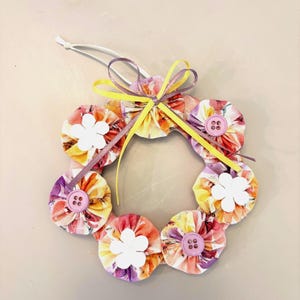 May include: A handmade fabric wreath with a yellow ribbon and pink buttons. The wreath is made of fabric flowers in shades of pink, purple, and yellow.