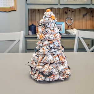 May include: A white and orange fabric cone-shaped tree with black accents and orange star decorations. The tree is decorated with black bats and is sitting on a gray table.