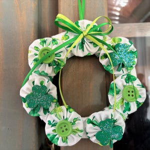 May include: A St. Patrick's Day wreath featuring white fabric circles with green shamrock patterns, green buttons, and glitter shamrock accents. The wreath is adorned with a green and yellow ribbon bow and a green hanging ribbon.