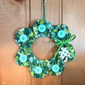 May include: A handmade St. Patrick's Day wreath. The wreath is made of green fabric flowers with light blue buttons in the center. A green ribbon and a shamrock accent the wreath. The wreath hangs from a green ribbon.