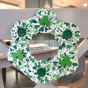 May include: A St. Patrick's Day wreath featuring a circular design with white fabric flowers adorned with green shamrock patterns and green buttons. Wooden shamrock cutouts are attached to the wreath. The wreath is displayed on a gray chair.