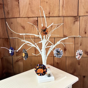 May include: A white birch tree with a white base, decorated with six fabric ornaments in orange, black, purple, and white. The ornaments are hanging from the branches of the tree. A small white owl figurine is sitting on the base of the tree.