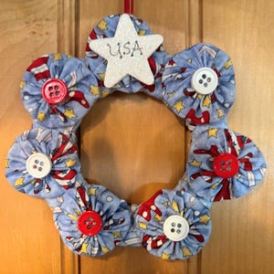 May include: A blue and red fabric wreath with a white star in the center that says "USA". The wreath is decorated with red and white buttons.