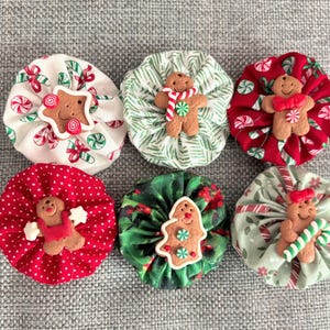 May include: Six handmade fabric ornaments in the shape of a flower with a gingerbread man in the center. The ornaments are in red, green, and white with candy cane and holly designs.