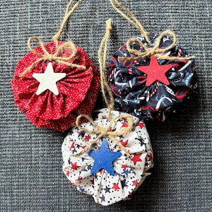 May include: Three patriotic fabric ornaments with star accents and twine hangers. One is red with white polka dots and a white star. The second is navy with red and white patterns and a red star. The third is white with red and blue stars and a blue star.