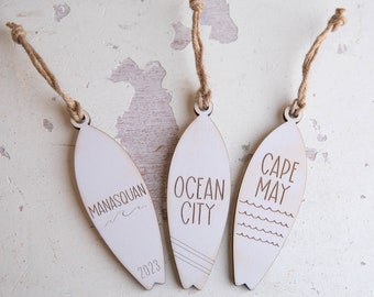 May include: Three wooden surfboard ornaments with twine hangers. The ornaments are engraved with the names of beach towns: Manasquan, Ocean City, and Cape May. The Manasquan ornament also includes the year 2023.