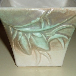 May include: A square ceramic planter with a light green glaze and a raised design of leaves and flowers. The planter has a white base and a light brown glaze on the top.
