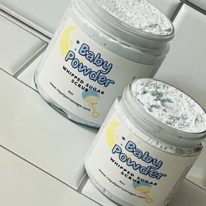 May include: Two jars of whipped sugar scrub with a light blue base and white whipped cream texture. The labels are white with blue text that reads "Baby Powder Whipped Sugar Scrub" and the website address "www.royalpersonage.com". The jars are on a white tiled surface.