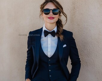 May include: A person wearing a dark blue suit with a white shirt and a black bow tie. They are wearing sunglasses and have a red lipstick. The person is standing in front of a beige wall.