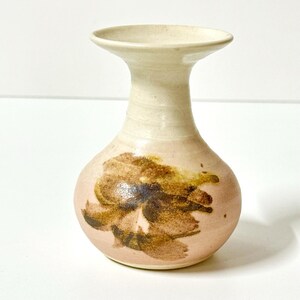 May include: A ceramic vase with a bulbous base and a flared rim. The piece features a light cream and pale pink glaze with a prominent, abstract brown and dark ochre brushstroke design centred on the front of the rounded body.