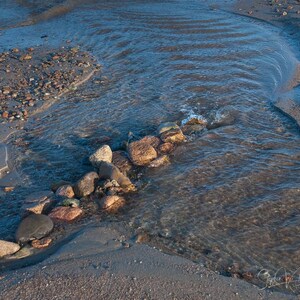 May include: A close-up of a small stream of water flowing over smooth, rounded rocks on a sandy beach. The water is clear and shallow, and the rocks are a variety of colors, including brown, gray, and red.