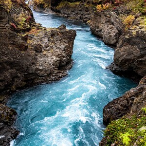 Puede incluir: Un paisaje vibrante con un río turquesa que fluye a través de un cañón rocoso. El agua cae sobre las rocas, creando rápidos blancos. Las paredes del cañón son de color marrón oscuro con parches de vegetación verde y naranja.