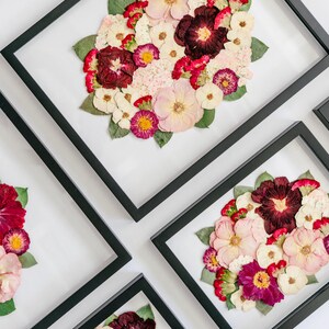 May include: A collection of rectangular black frames containing pressed flower arrangements. Each frame displays a variety of dried blossoms in shades of deep burgundy, soft pink, cream, and white, accented with green leaves on a white background.