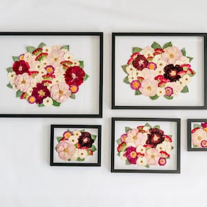 May include: Five rectangular black frames containing pressed flower arrangements. Each frame displays a cluster of dried blossoms in shades of deep red, light pink, and white, accented with green leaves, all set against a clear glass background.