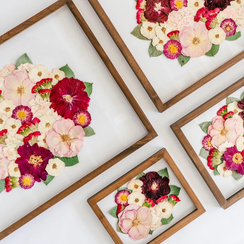 May include: Four rectangular wooden frames containing pressed flower arrangements. Each frame displays a collection of dried blossoms in shades of deep red, magenta, soft pink, and white, accented with green leaves, all set against a white background.
