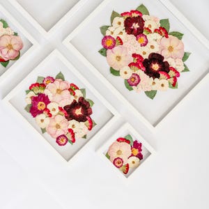 May include: Three white rectangular frames containing pressed flower arrangements. The flowers feature shades of pink, deep red, burgundy, and white, accented with green leaves. The compositions are displayed against a white background.