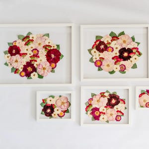 May include: Five white rectangular floating frames containing pressed flower arrangements. Each frame displays a cluster of dried flowers in shades of deep burgundy, soft pink, cream, and white, accented with small green leaves, arranged in a circular pattern.