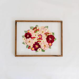 May include: A rectangular wooden frame containing a pressed floral arrangement. The composition features a cluster of dried flowers in shades of deep burgundy, soft pink, and white, accented with small green leaves against a white background.