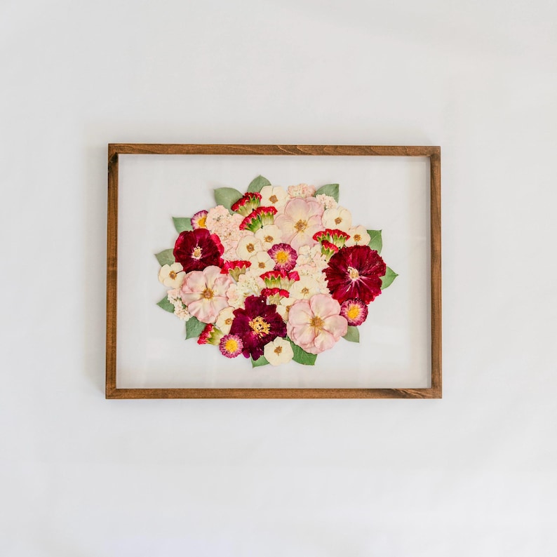 May include: A rectangular wooden frame containing a pressed floral arrangement. The composition features a cluster of dried flowers in shades of deep burgundy, soft pink, and white, accented with small green leaves against a white background.