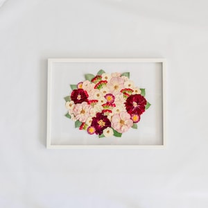 May include: A rectangular white frame containing a pressed flower arrangement. The bouquet features a mix of deep burgundy, soft pink, and white blossoms interspersed with small green leaves, all displayed against a clear glass background.