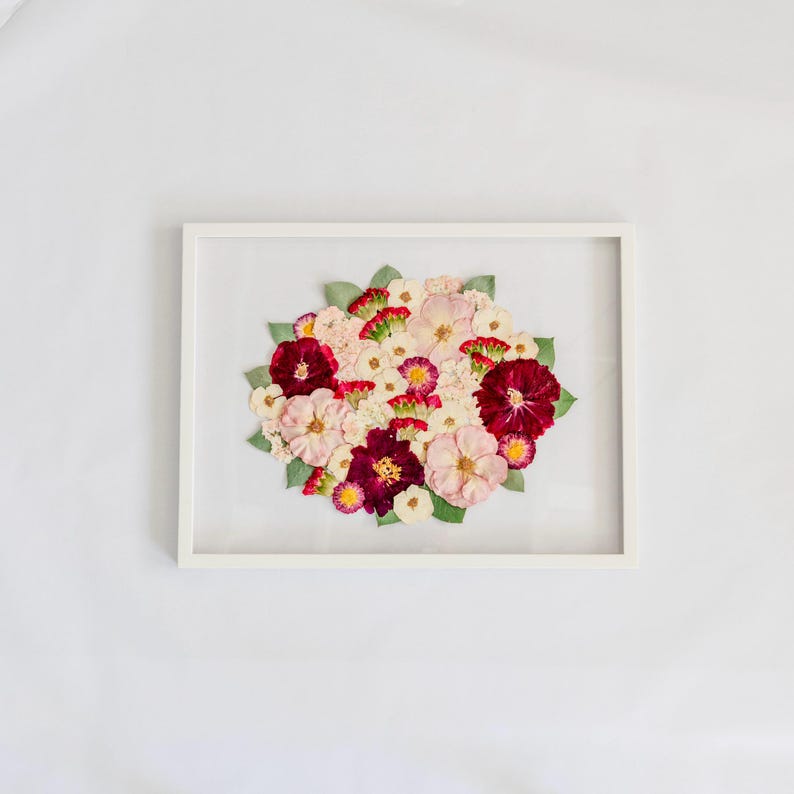 May include: A rectangular white frame containing a pressed flower arrangement. The bouquet features a mix of deep burgundy, soft pink, and white blossoms interspersed with small green leaves, all displayed against a clear glass background.