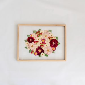 May include: A rectangular light wood frame containing a pressed flower arrangement. The bouquet features a mix of deep burgundy, soft pink, and white blossoms interspersed with small green leaves, all displayed against a white background.