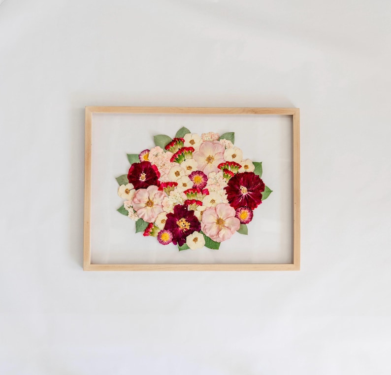 May include: A rectangular light wood frame containing a pressed flower arrangement. The bouquet features a mix of deep burgundy, soft pink, and white blossoms interspersed with small green leaves, all displayed against a white background.