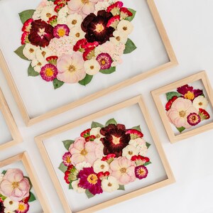 May include: A collection of four light-colored wooden frames containing pressed flower arrangements. The displays feature a variety of dried blossoms in shades of deep burgundy, soft pink, cream, and white, accented with green leaves on a clear background.